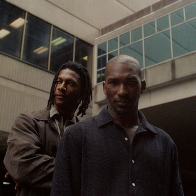 Two men pose in front of a concrete building.